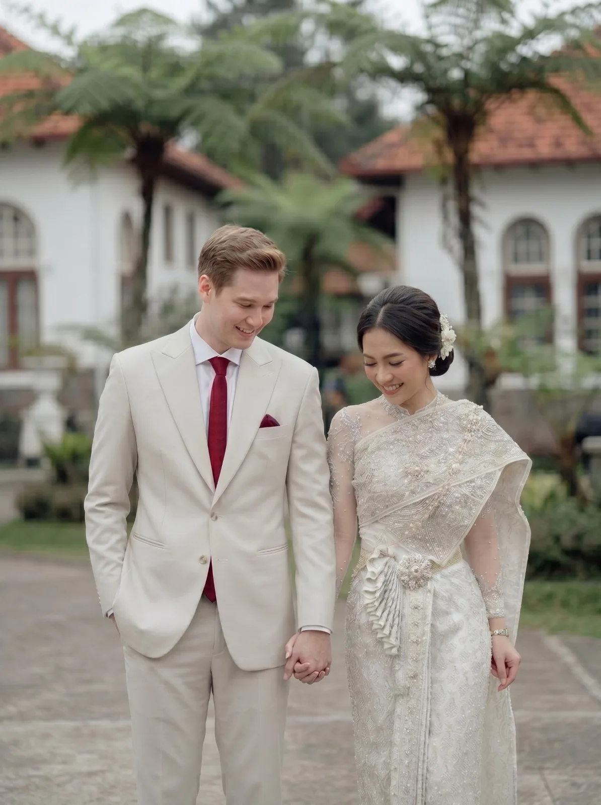 Pengantin pria mengenakan jas pernikahan bespoke beige dengan dasi burgundy oleh Laxmi Tailors Surabaya, potret bersama pengantin wanita